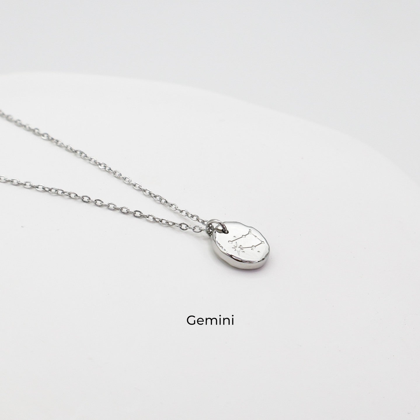 Silver necklace with a round pendant on a light gray background. The pendant is engraved with the Gemini star sign constellation