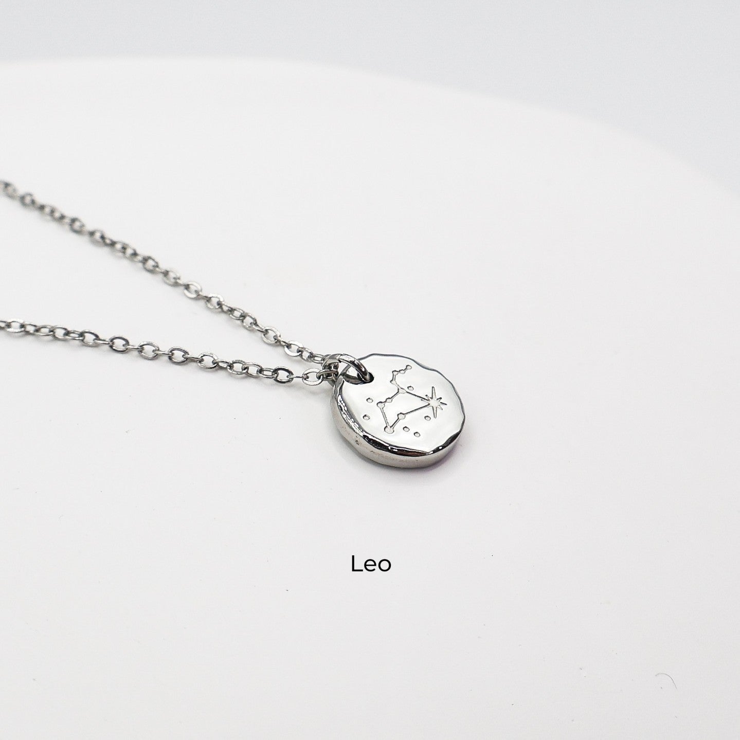 Silver necklace with a round pendant on a light gray background. The pendant is engraved with the Leo star sign constellation