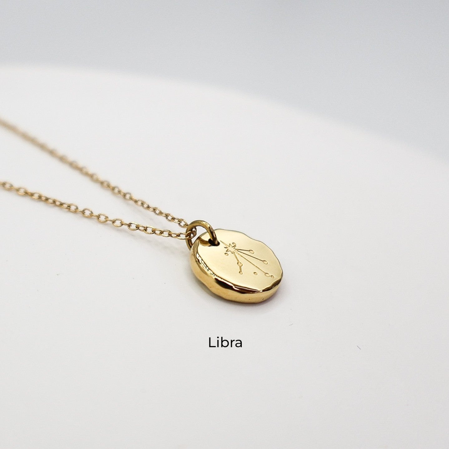Gold necklace with a round pendant on a light gray background. The pendant is engraved with the Libra star sign constellation